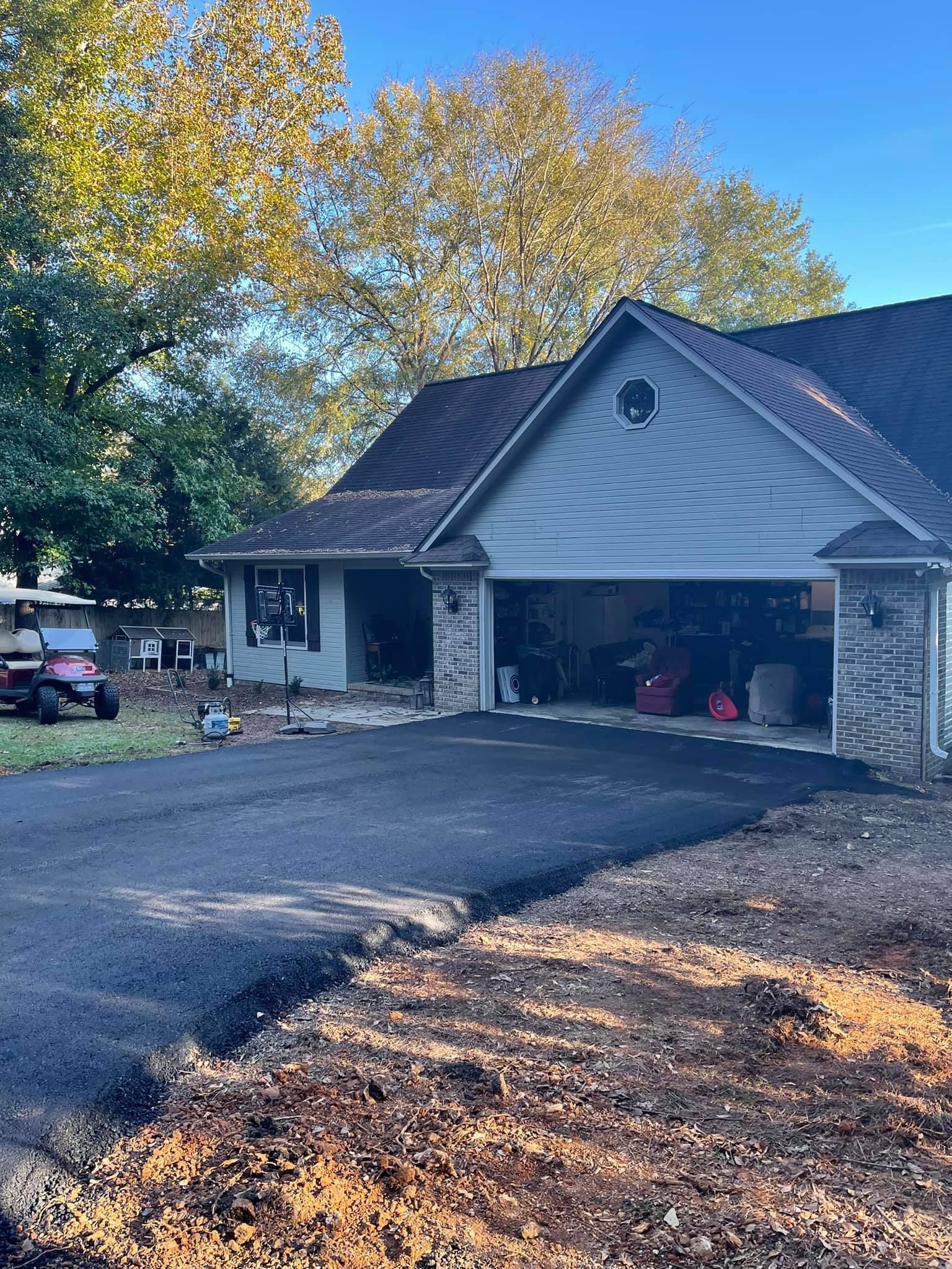 A house with a newly paved asphalt driveway, a garage, and trees with autumn foliage under a clear blue sky.