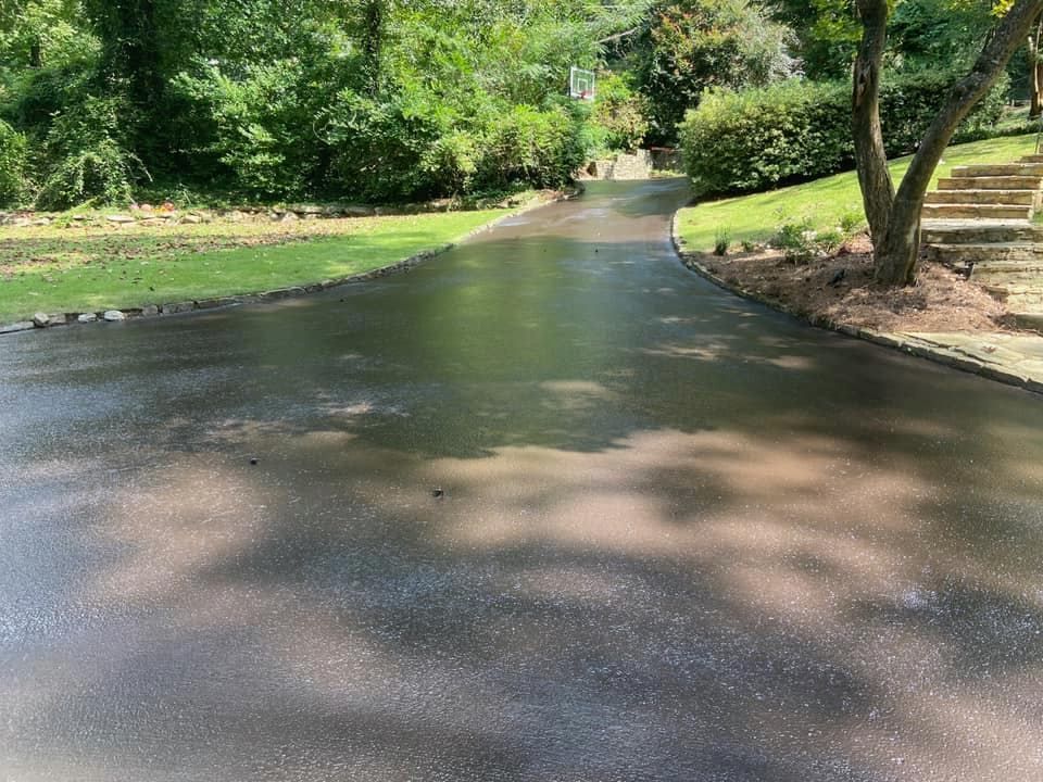 A wet, paved driveway curves through a lush green yard with trees and stone steps on a sunny day.