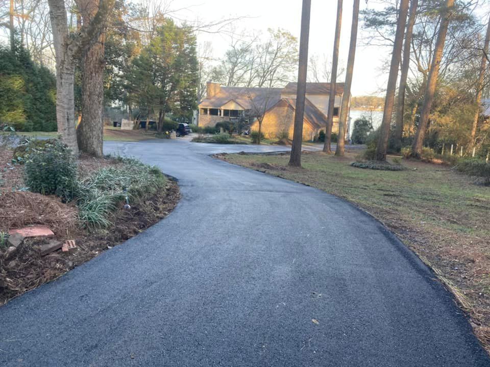 A winding asphalt driveway leads through a wooded yard toward a two-story house under a clear sky.