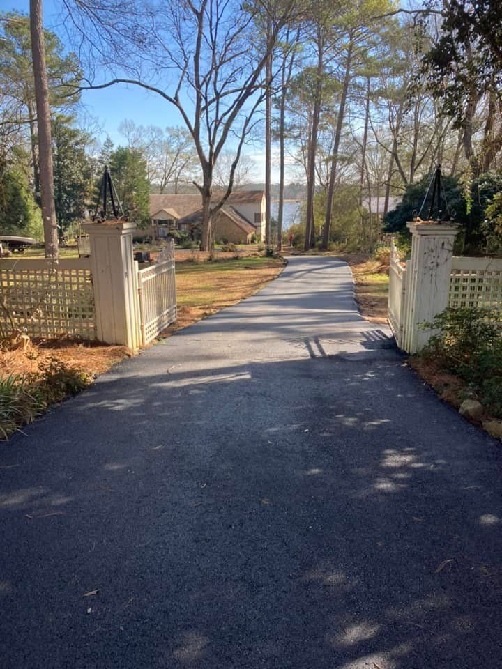 A paved driveway leads toward a house in the distance, framed by white gate pillars and a lattice fence under tall trees.
