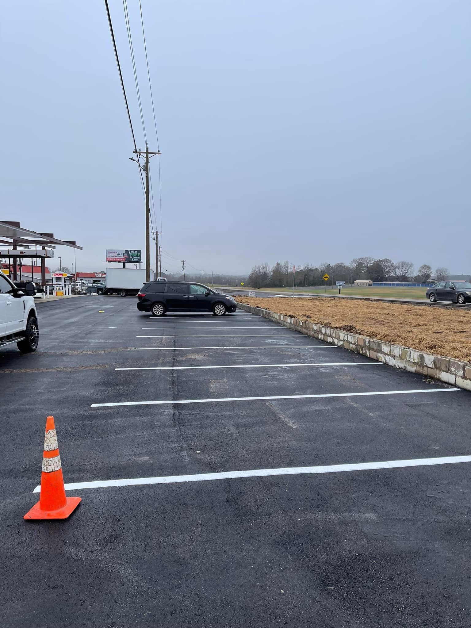A parking lot with freshly painted white parking lines, a black minivan, a traffic cone, and a dirt embankment.