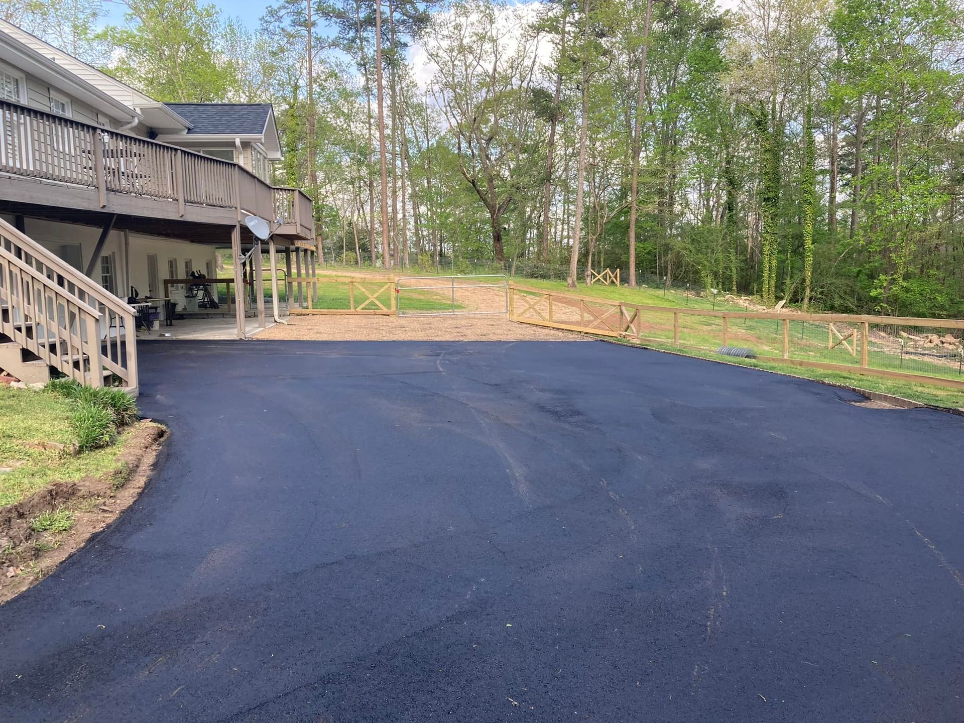 A paved driveway leads to the side of a two-story house with a wooden deck, bordered by a wooden fence and trees.