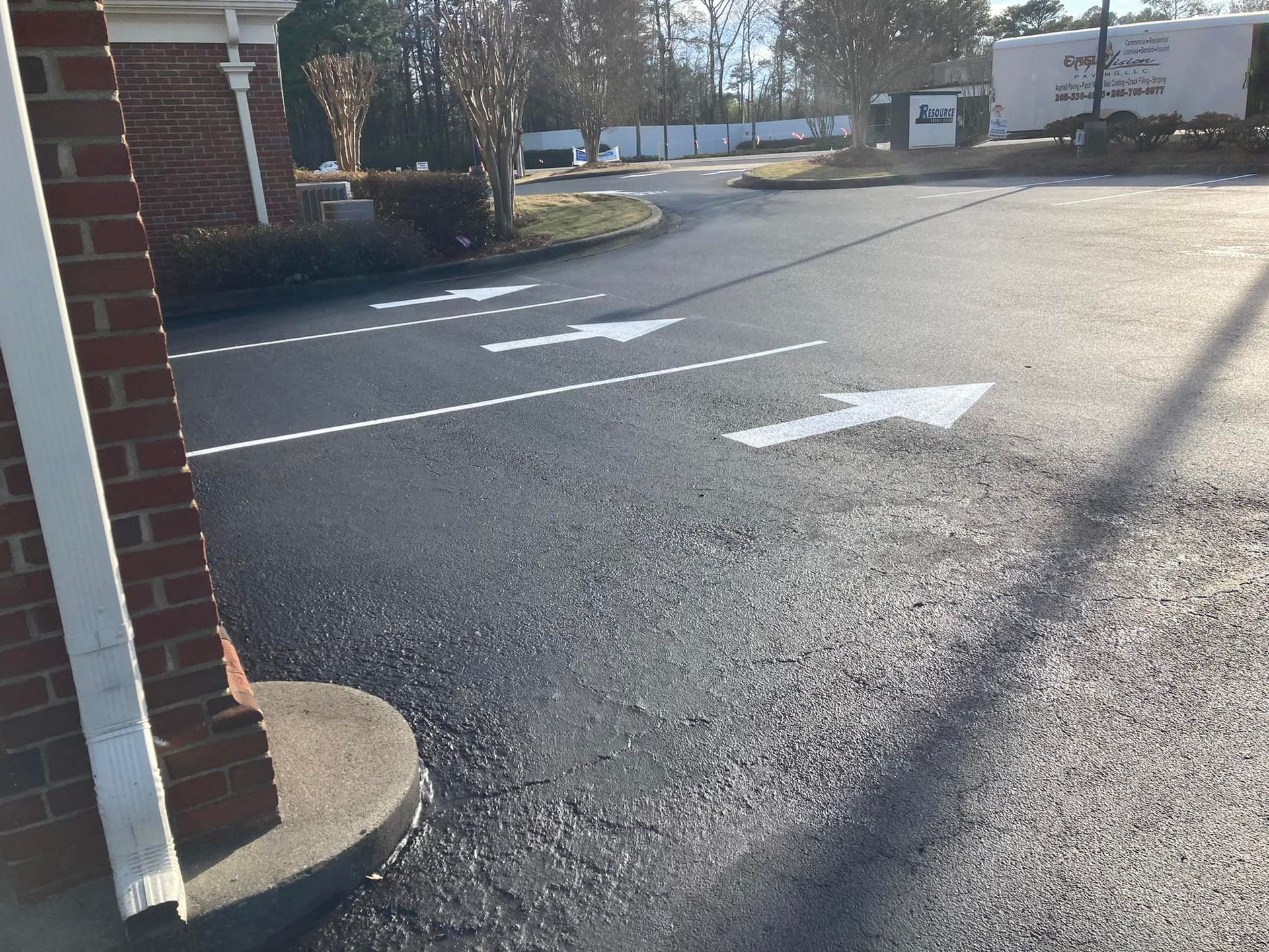 A paved drive-thru lane beside a brick building with white painted arrows indicating the direction of traffic flow.