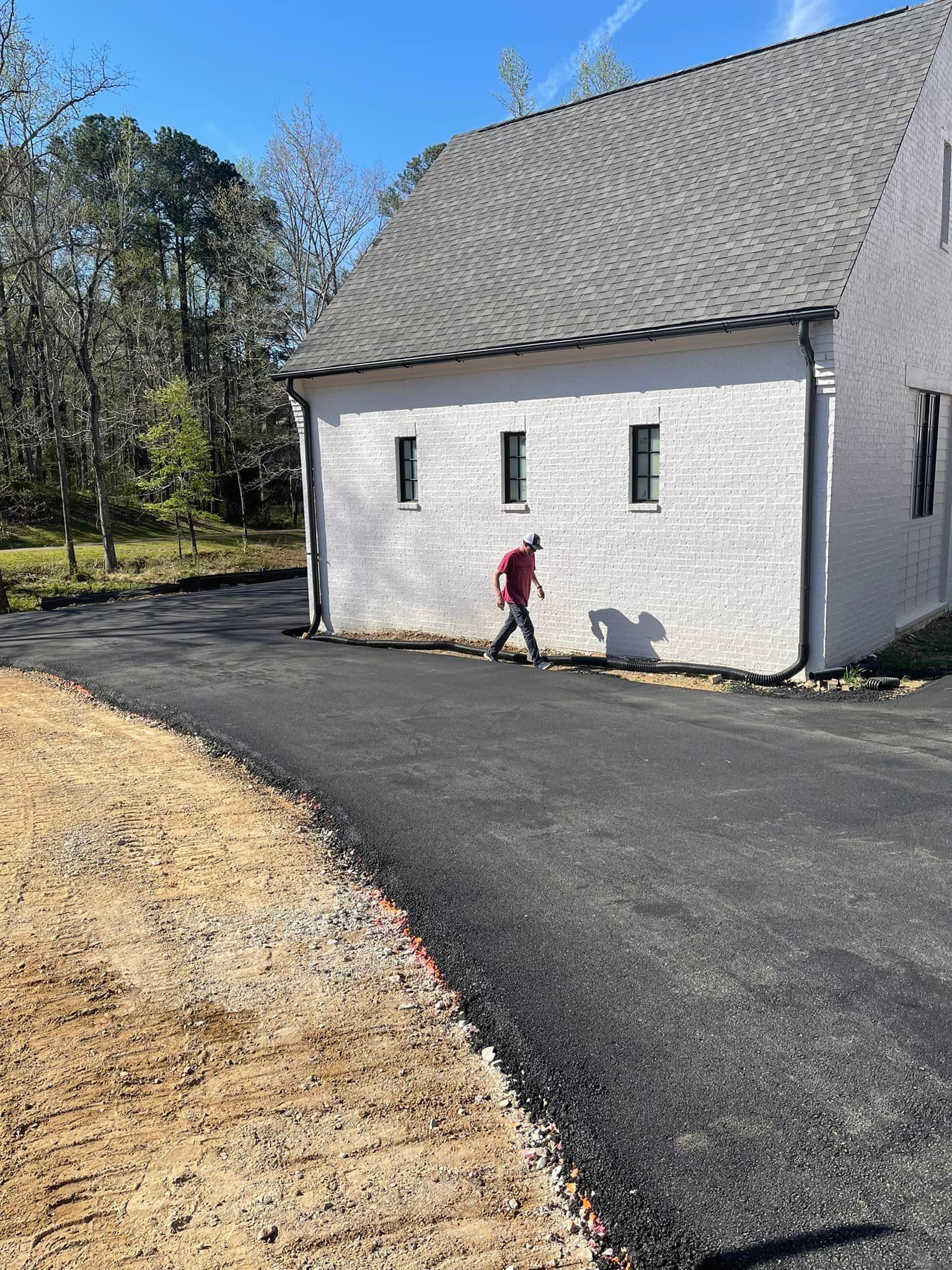 A person walks past a white brick building on a newly paved asphalt driveway next to a dirt landscape.