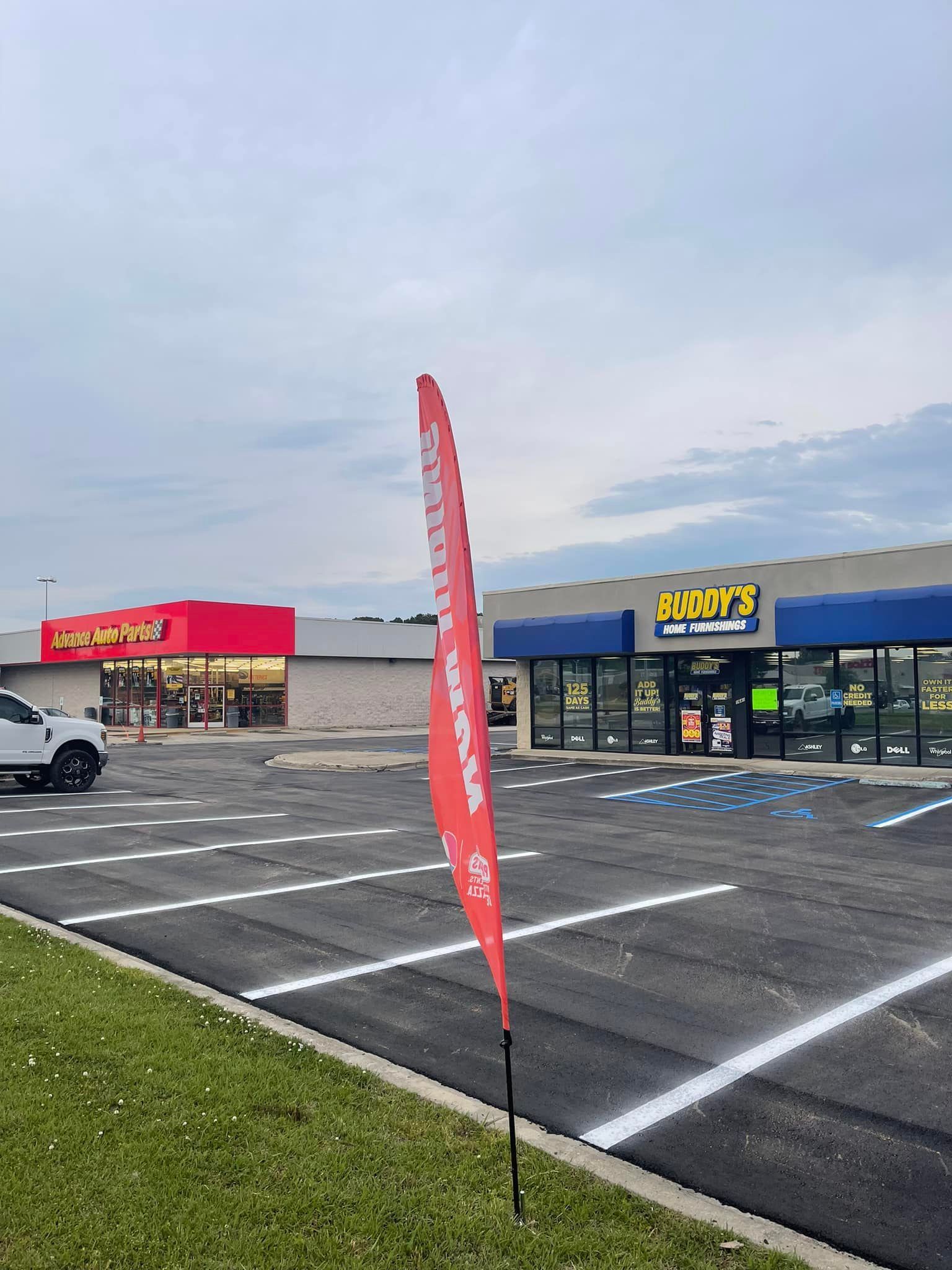 A parking lot in front of retail stores, including Buddy’s, with a red feather flag in the foreground.