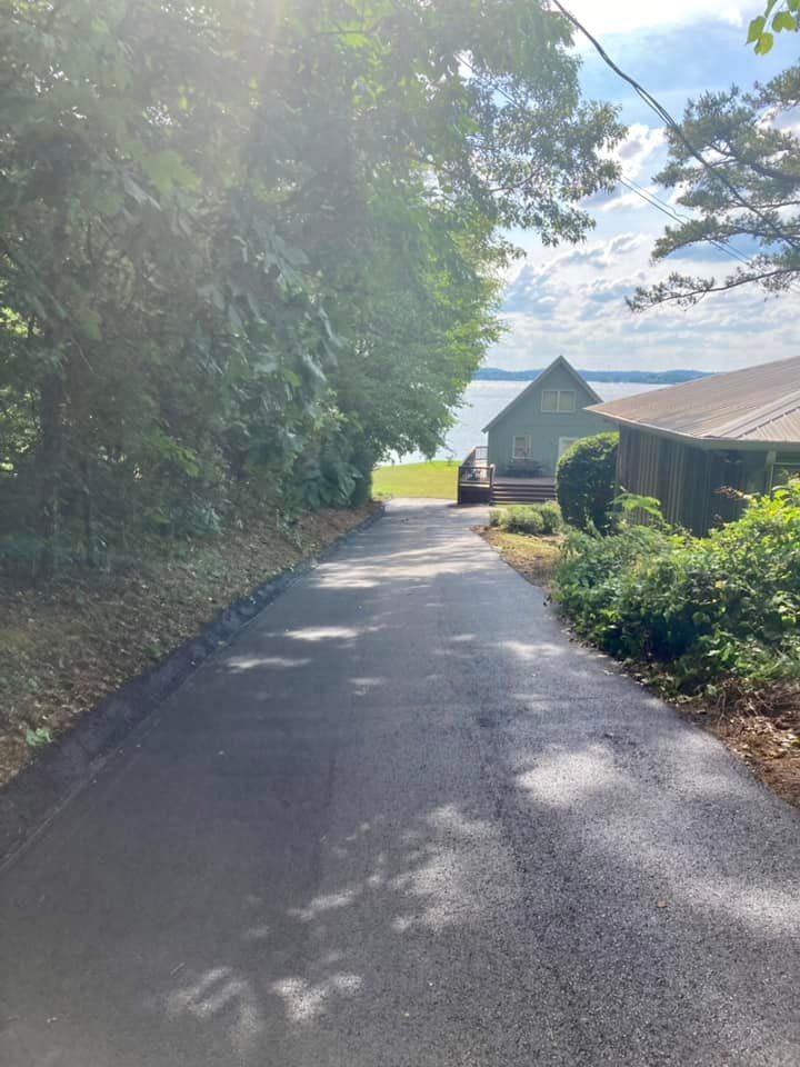 A steep, newly paved asphalt driveway leads down toward a lake, flanked by trees and two houses on the right.