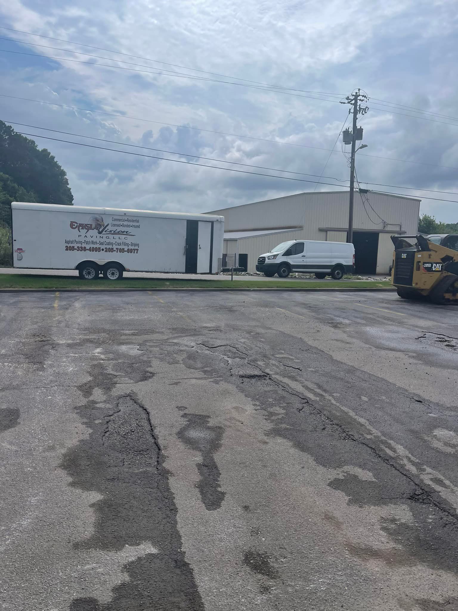 A white utility trailer parked near a white van and a construction vehicle in a gravel lot under a cloudy sky.