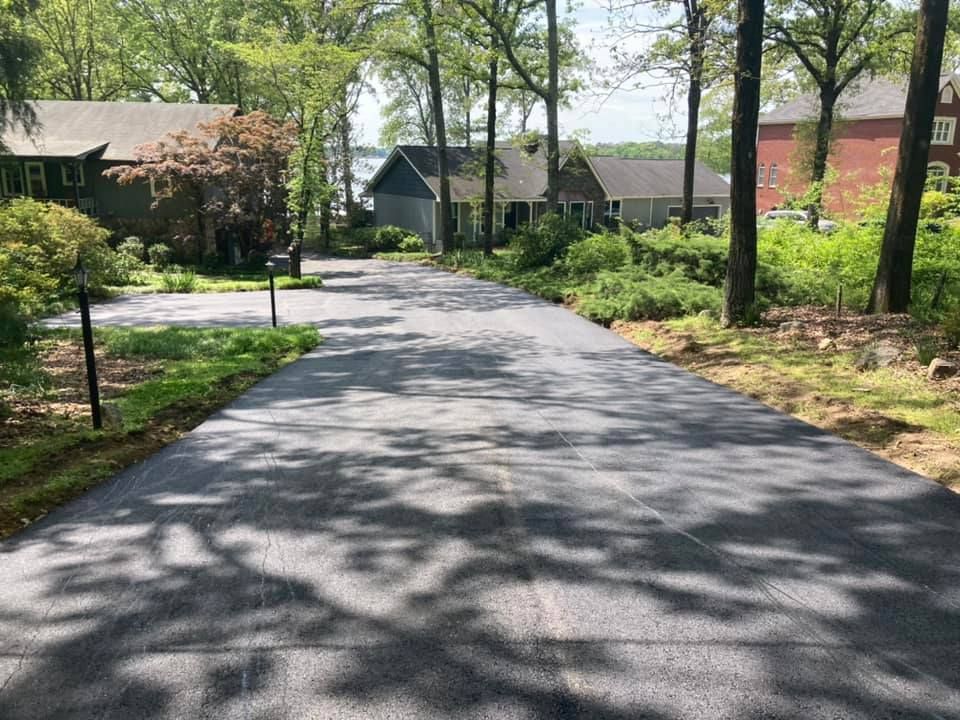 A newly paved asphalt driveway leads toward houses surrounded by lush green trees on a bright, sunny day.
