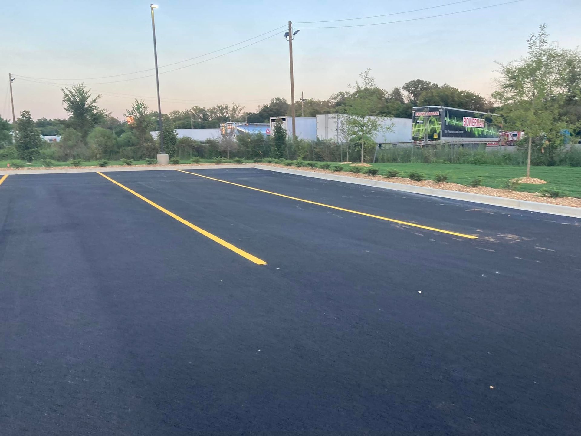 A newly paved parking lot with freshly painted yellow lines at dusk, bordered by trees and grassy areas.