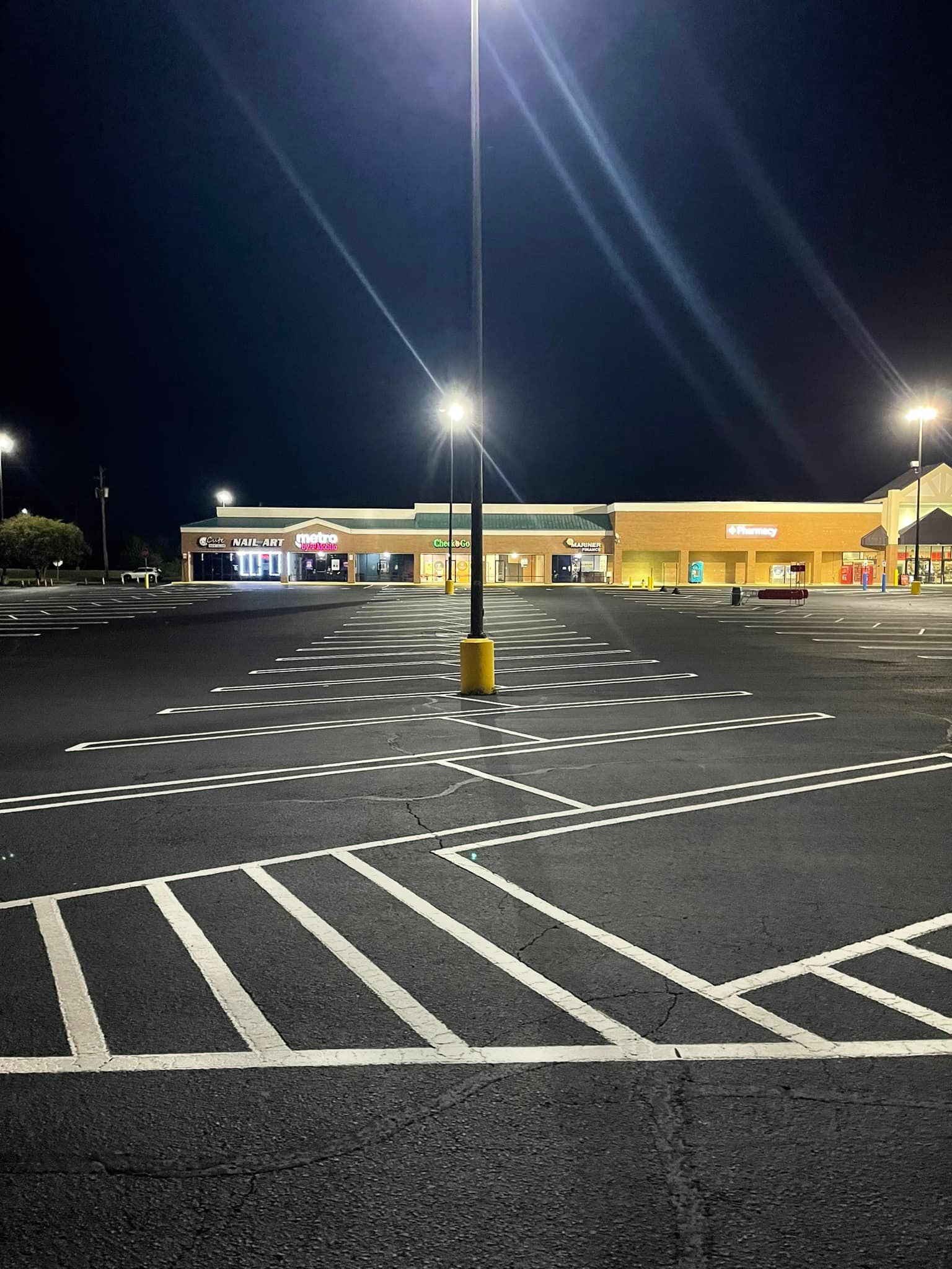 Empty parking lot at night with bright overhead lights illuminating the white lane markings and distant storefronts.
