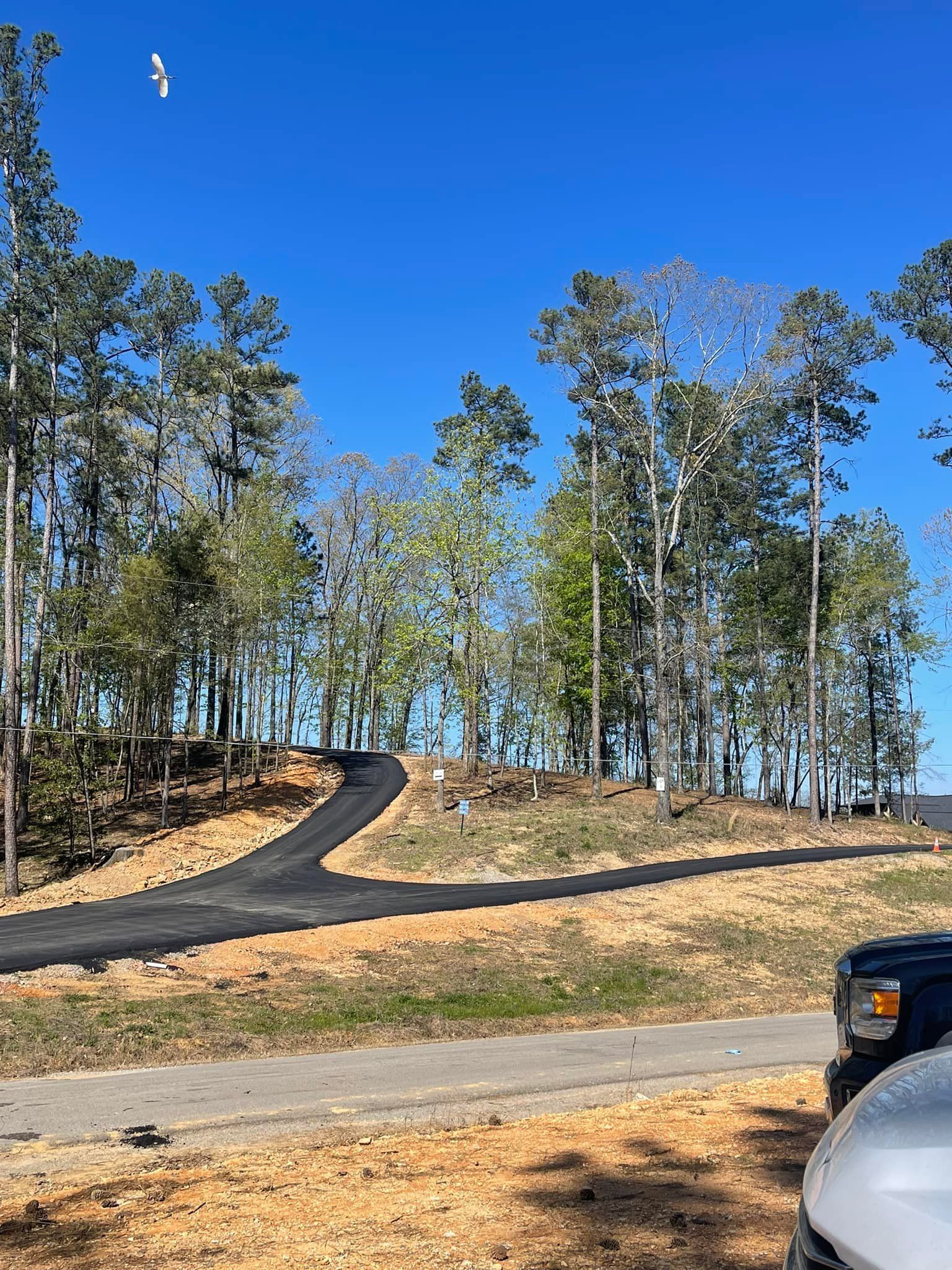 A freshly paved Y-shaped driveway leads uphill through a wooded area under a clear blue sky.