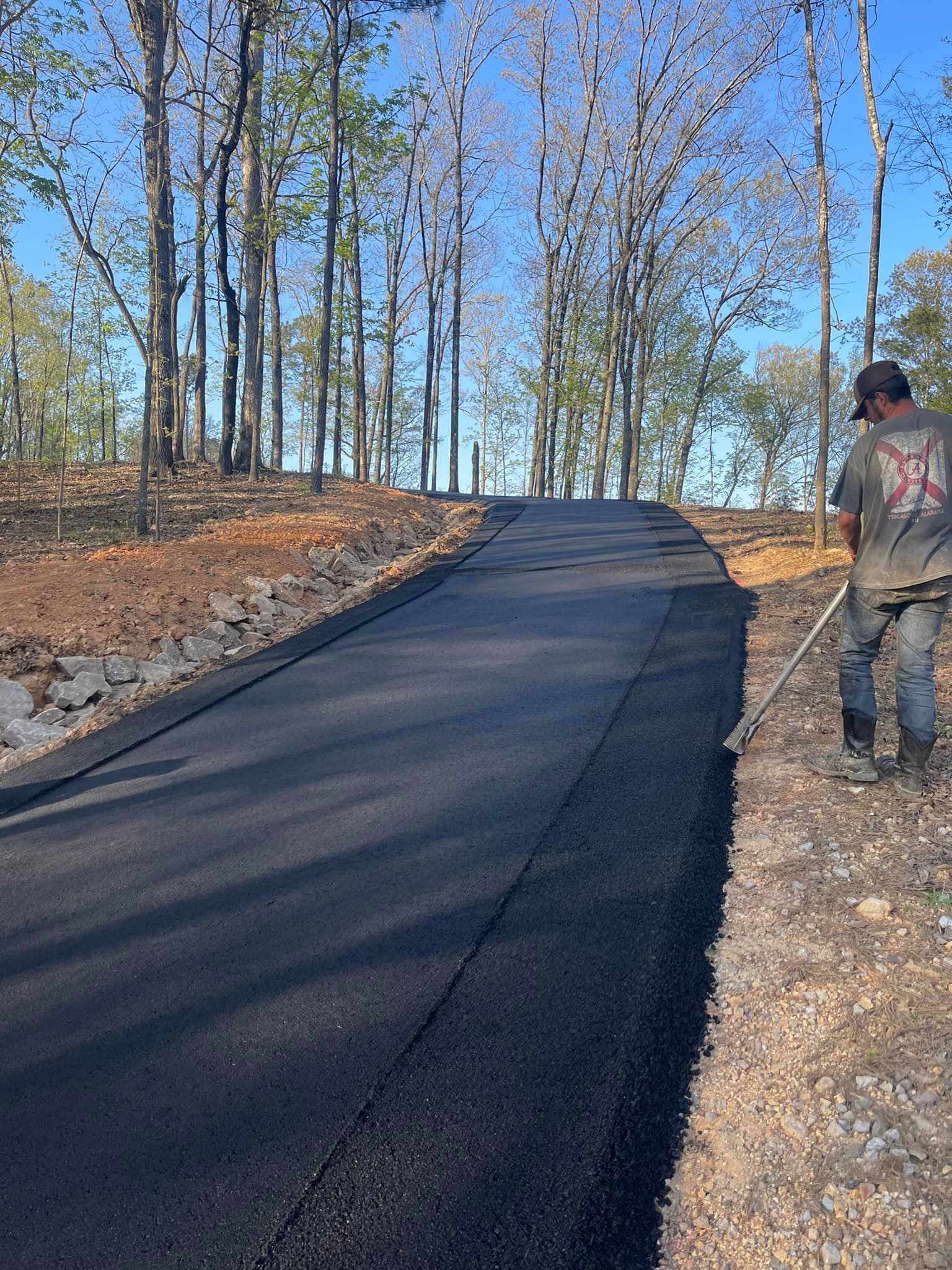 A worker in work clothes uses a hand tool to finish the edge of a newly paved asphalt driveway in a wooded area.
