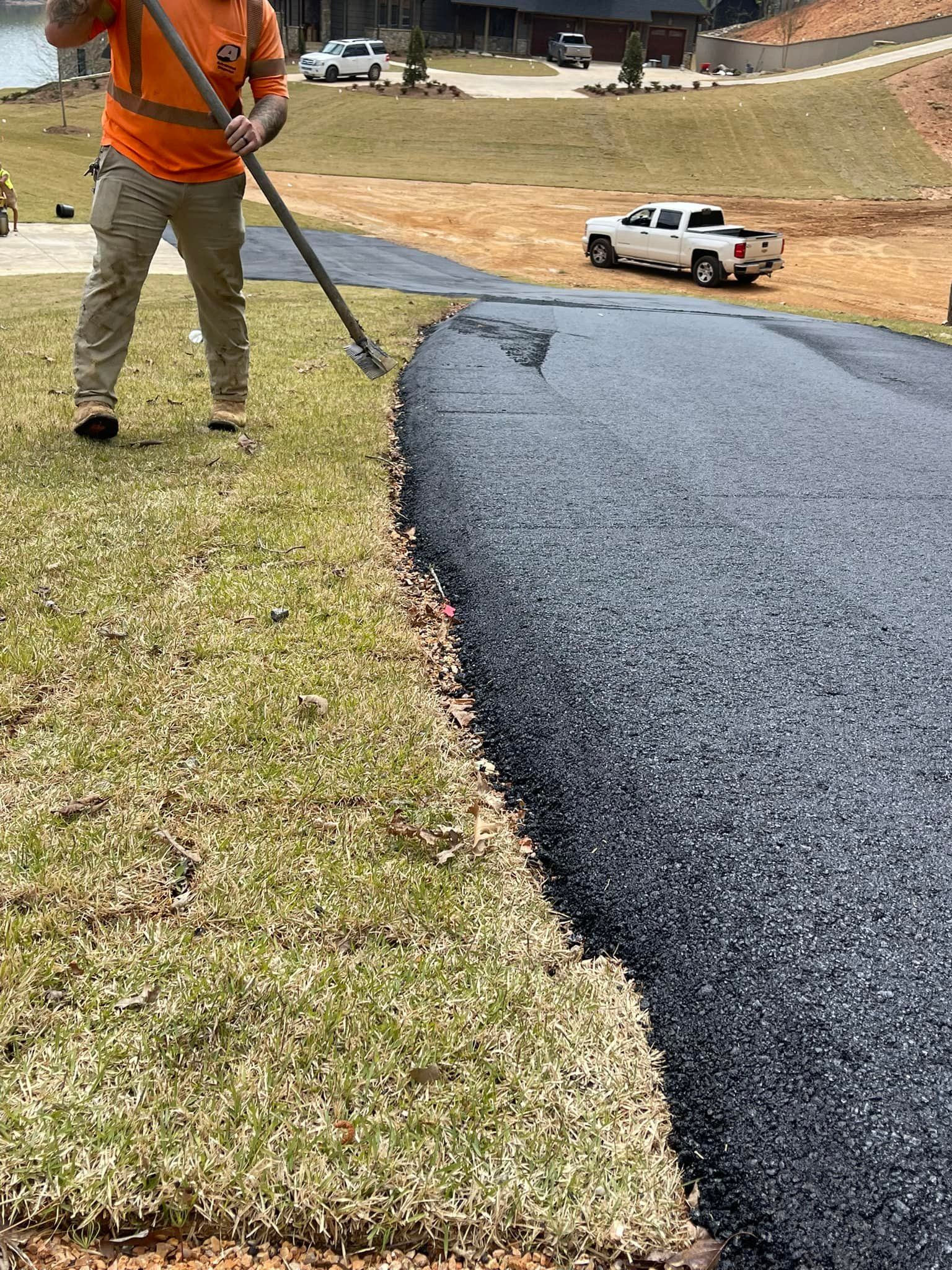 A worker in an orange shirt uses a tool to rake the edge of a freshly laid asphalt driveway on a sunny day.