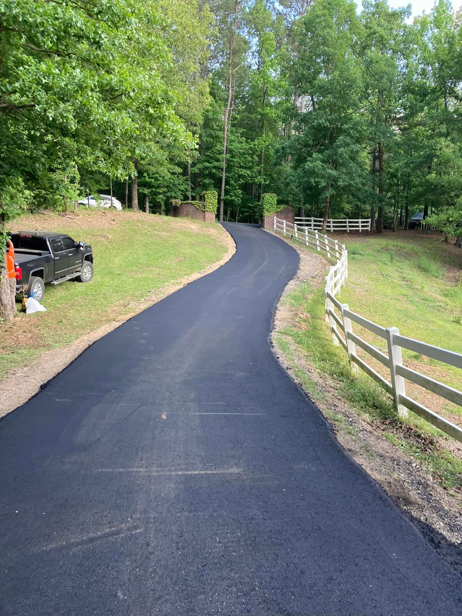 A curved, freshly paved asphalt driveway leads through a wooded area, bordered by a white rail fence and a parked truck.