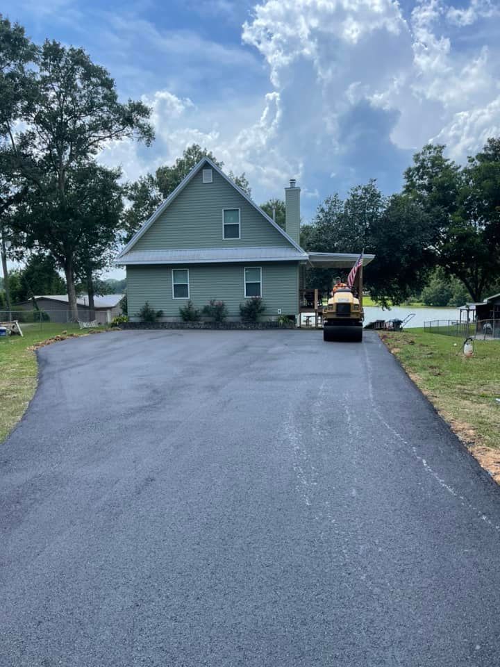 A freshly paved asphalt driveway leads to a light green house near a lake, with a steamroller parked in the foreground.