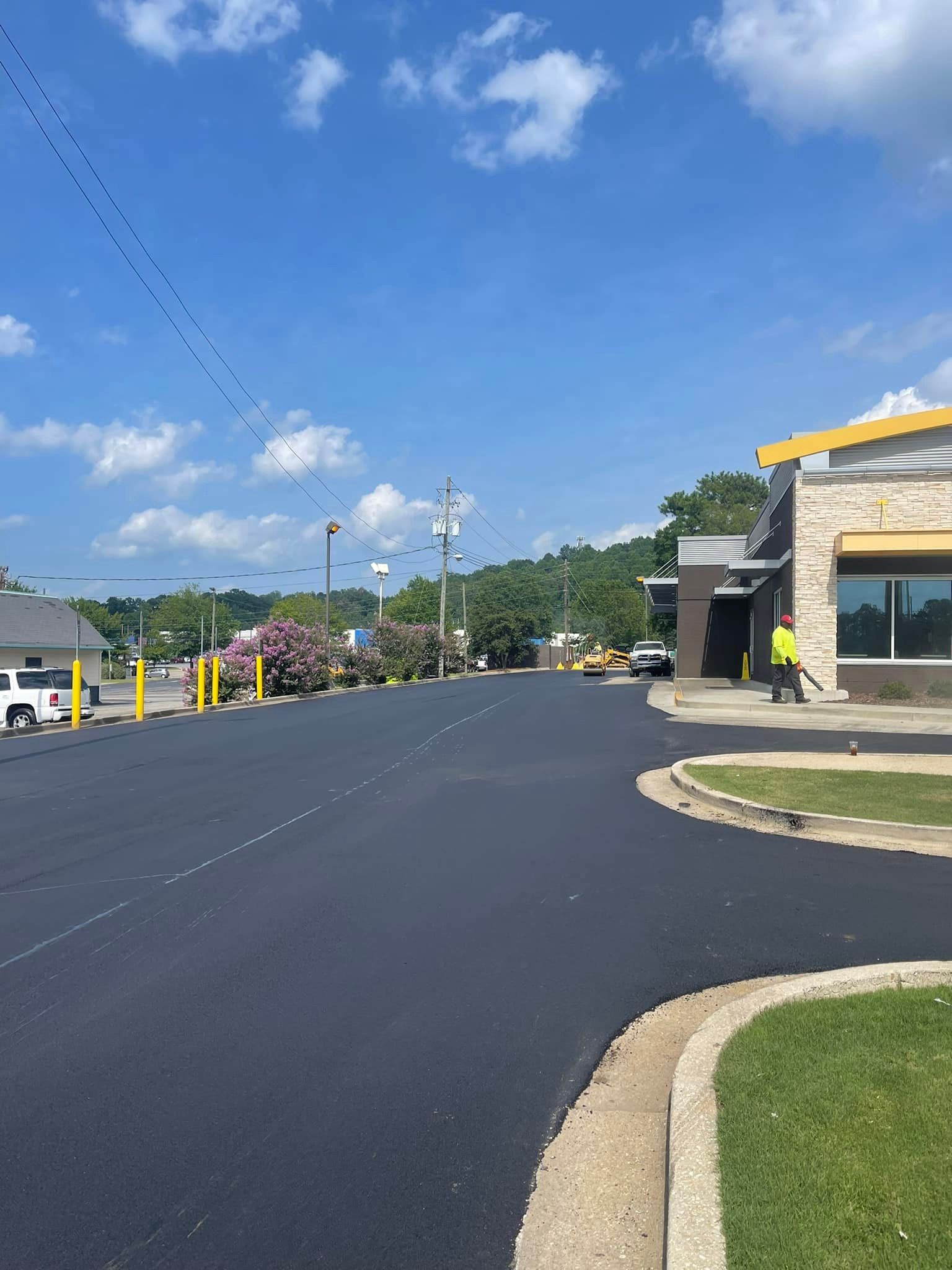 A freshly paved dark asphalt parking lot stretches toward a building under a bright blue sky with scattered clouds.