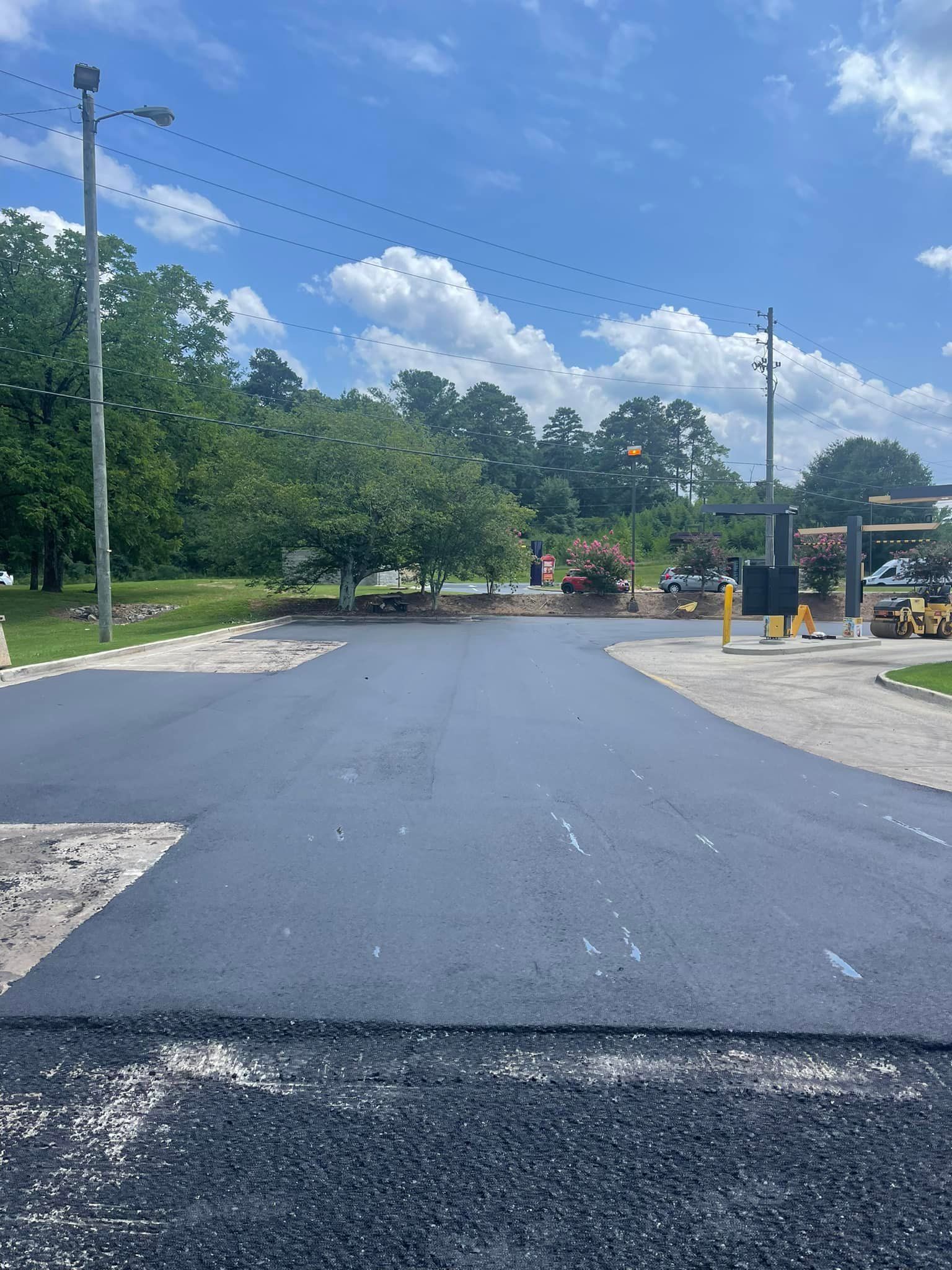 A paved parking lot with a freshly laid section of black asphalt, surrounded by older pavement and trees under a blue sky.