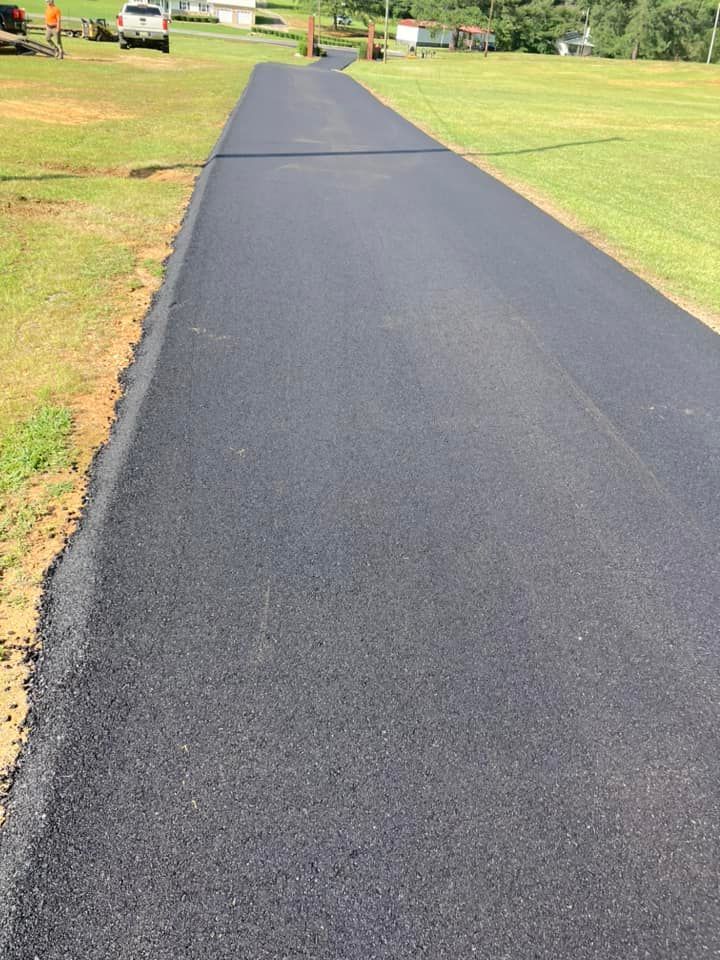 A newly paved asphalt driveway stretches through a green, grassy field toward a house in the distance.