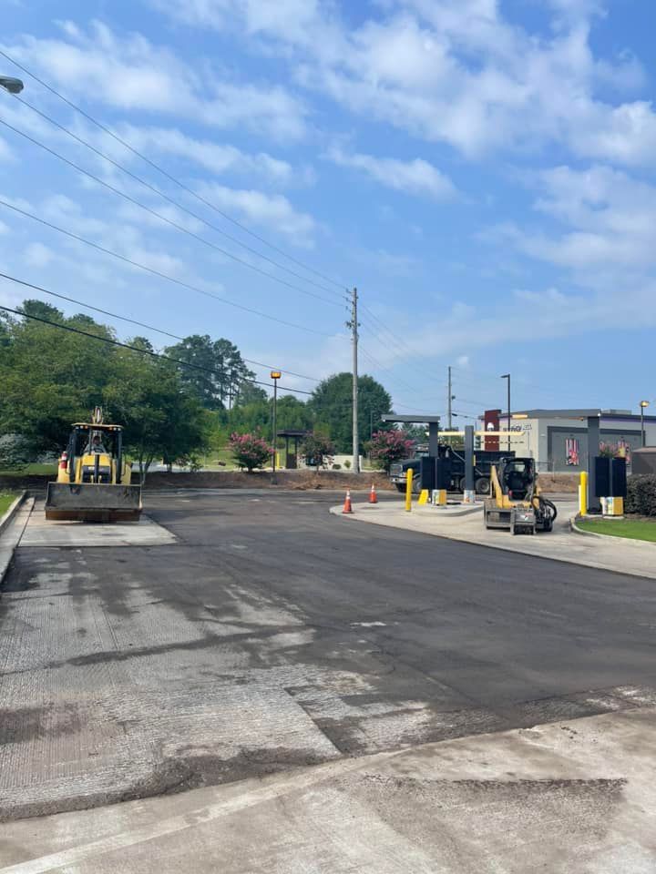 Construction equipment works on freshly paved asphalt in front of a building with a drive-thru lane under a clear sky.