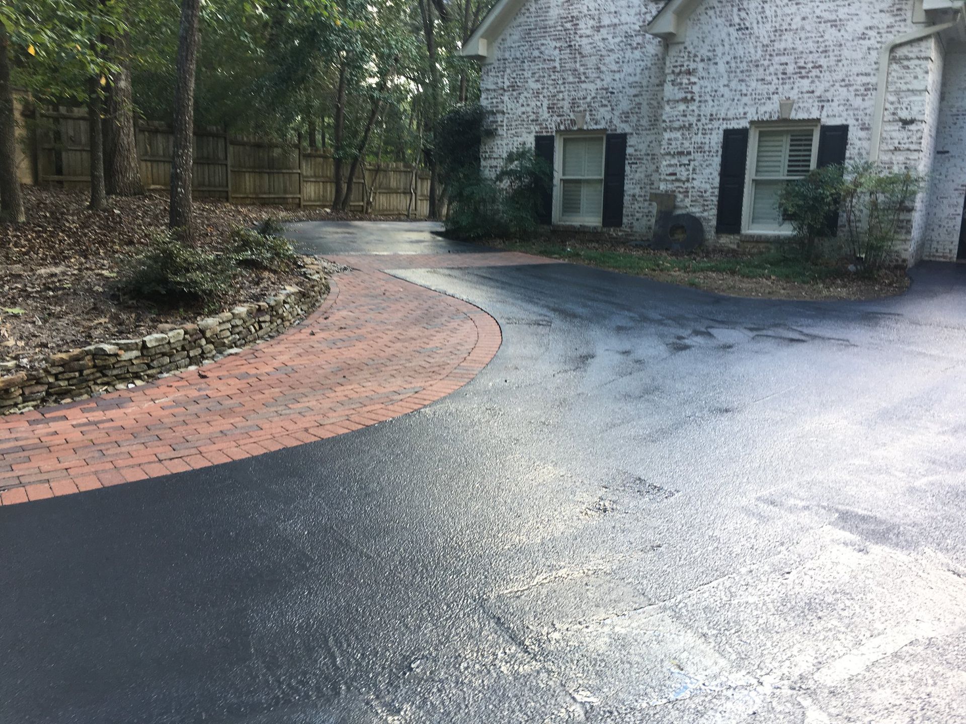A freshly sealed black asphalt driveway with a decorative curved border of red brick pavers next to a white brick house.