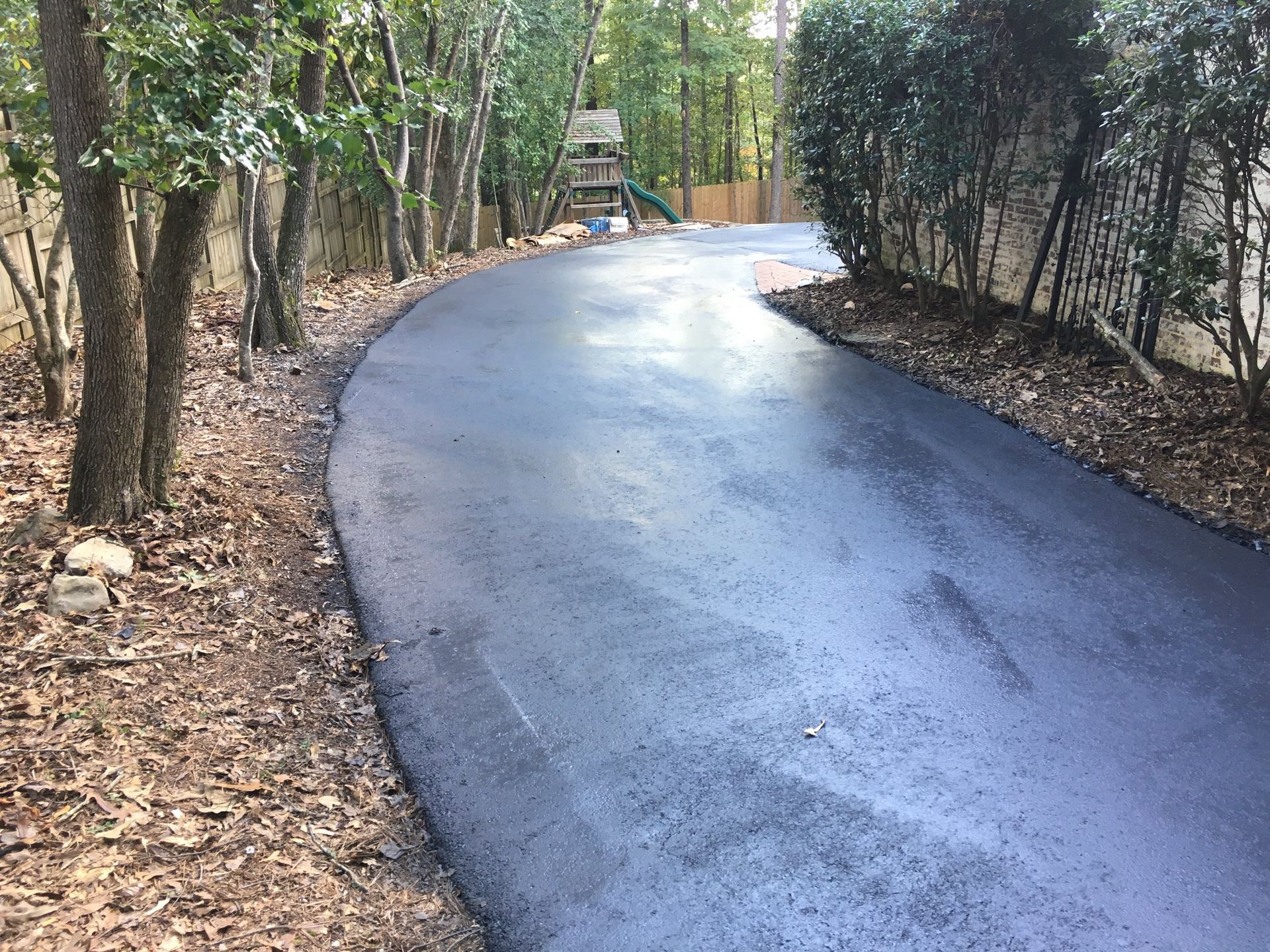 A freshly paved asphalt driveway curves through a wooded area lined with trees and a hedge.