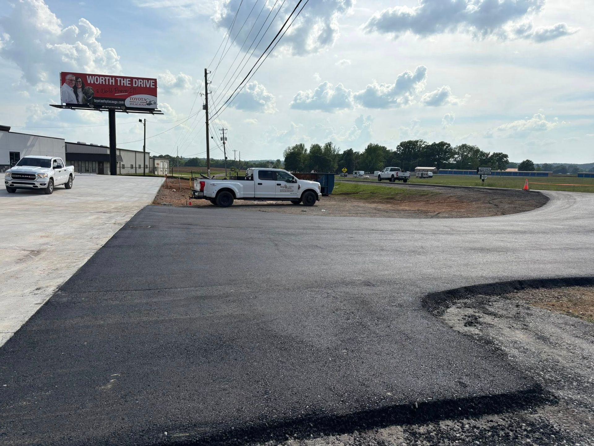 A freshly paved asphalt driveway leads to a parking area with white pickup trucks under a partly cloudy sky.