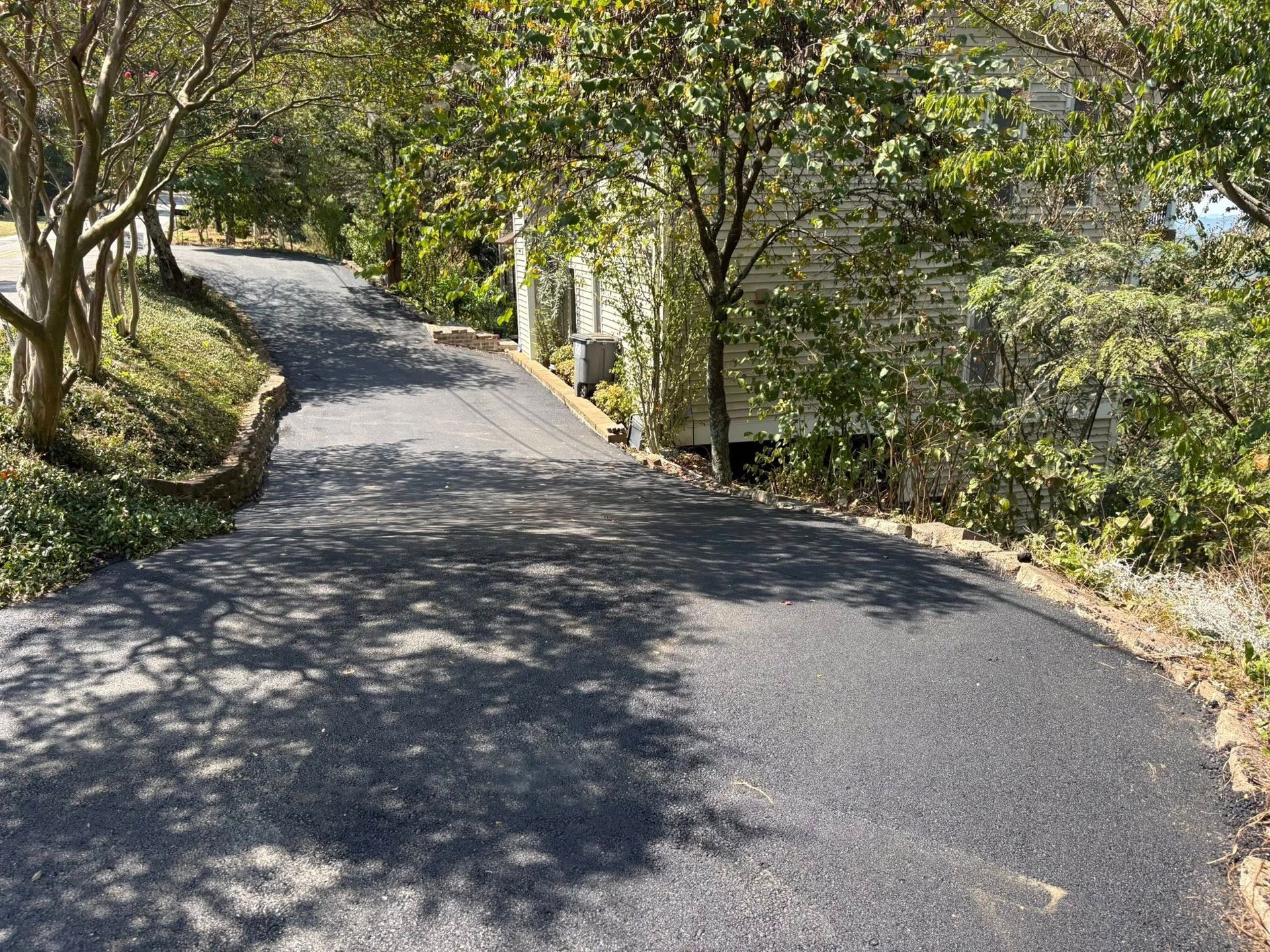 A freshly paved black asphalt driveway winds upward through a sunlit, tree-lined residential area.
