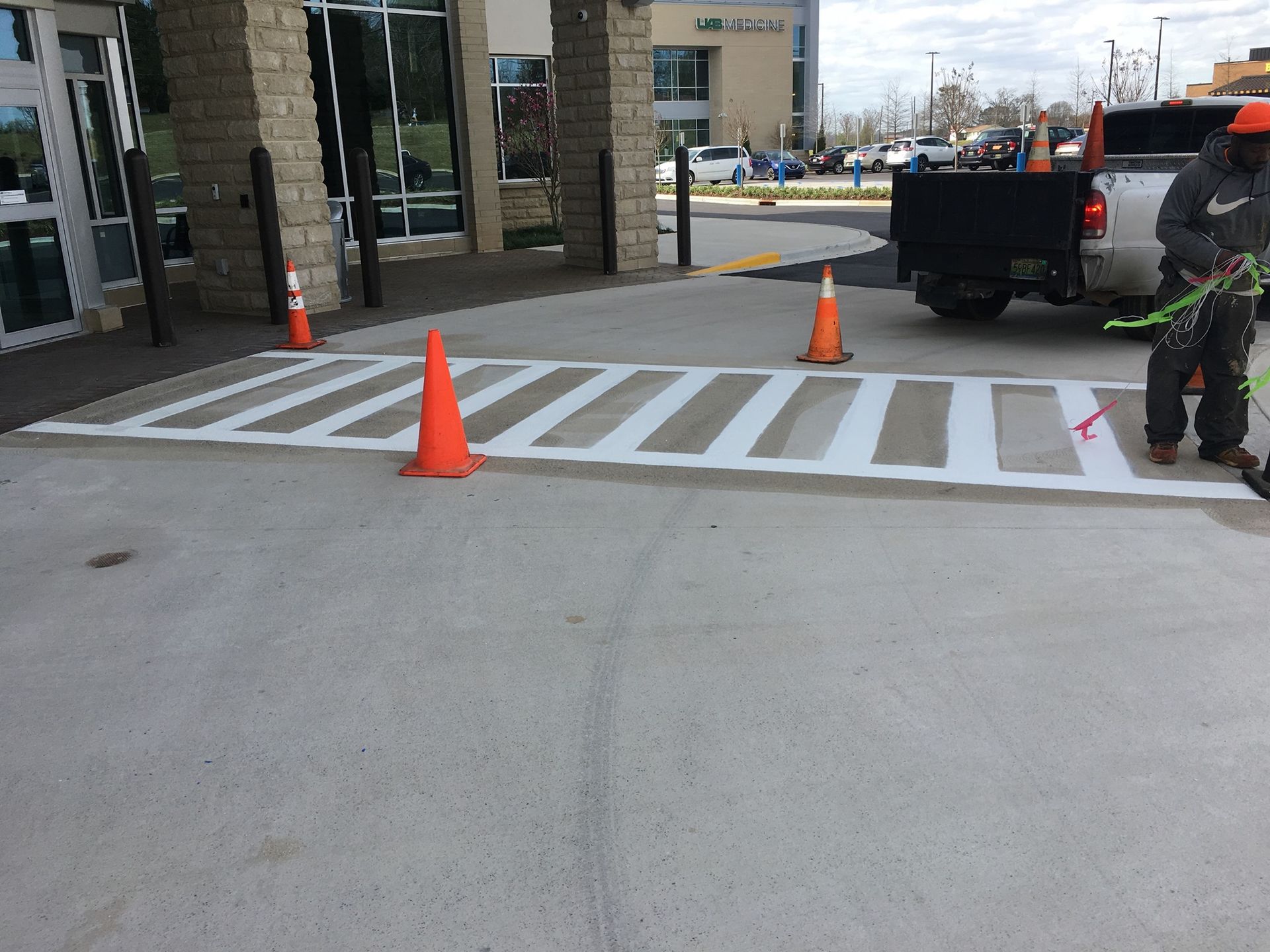 A worker paints a white crosswalk on concrete, marked by three orange safety cones in front of a building entrance.