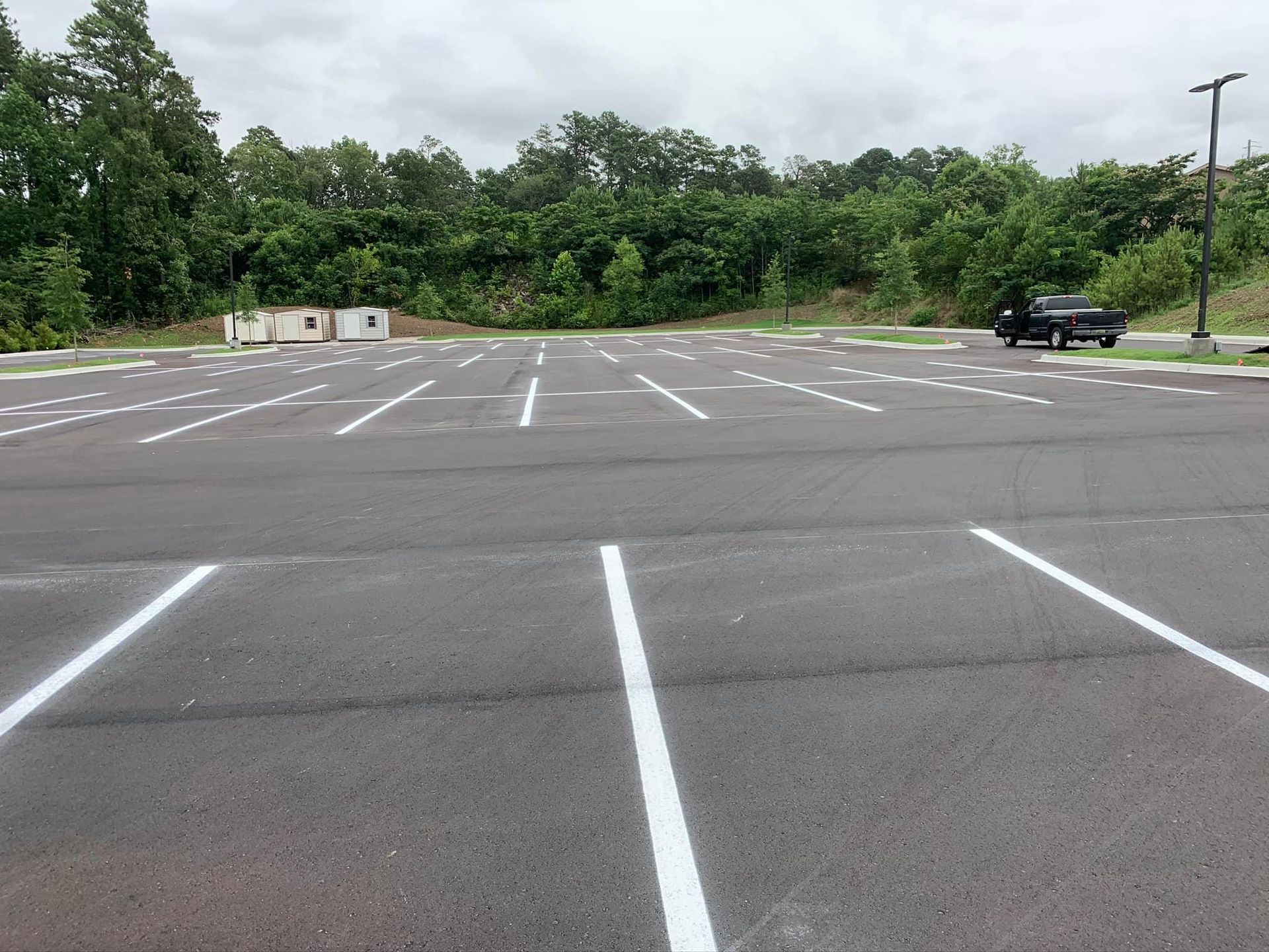 An empty asphalt parking lot with white painted stalls, bordered by a treeline and a single black pickup truck.