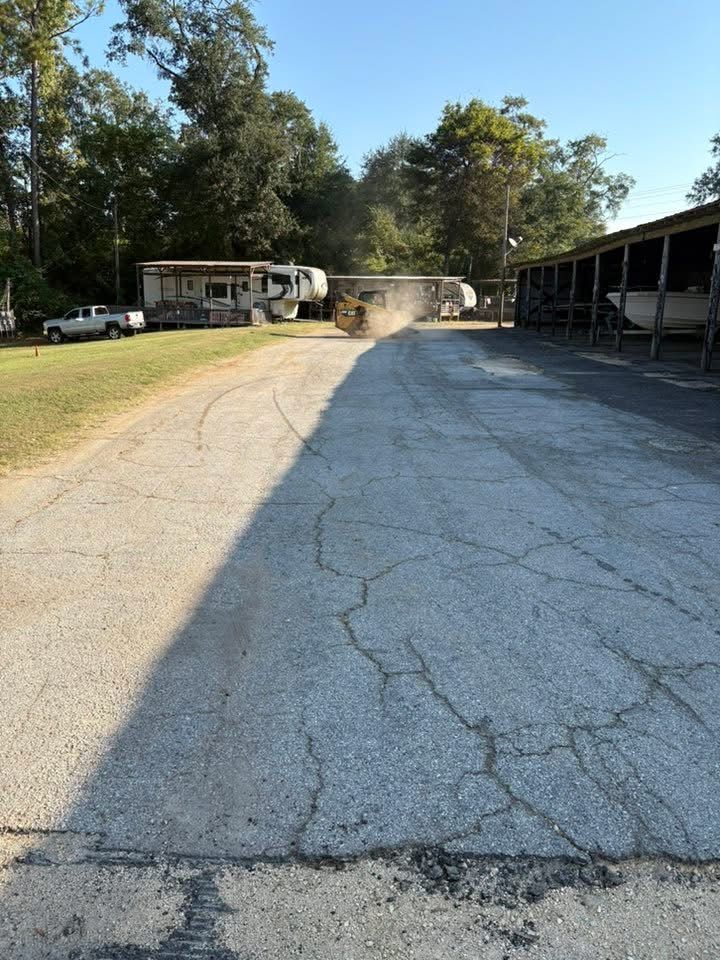 A view down a cracked asphalt driveway toward RVs and a boat shed, with a yellow piece of equipment stirring up dust.