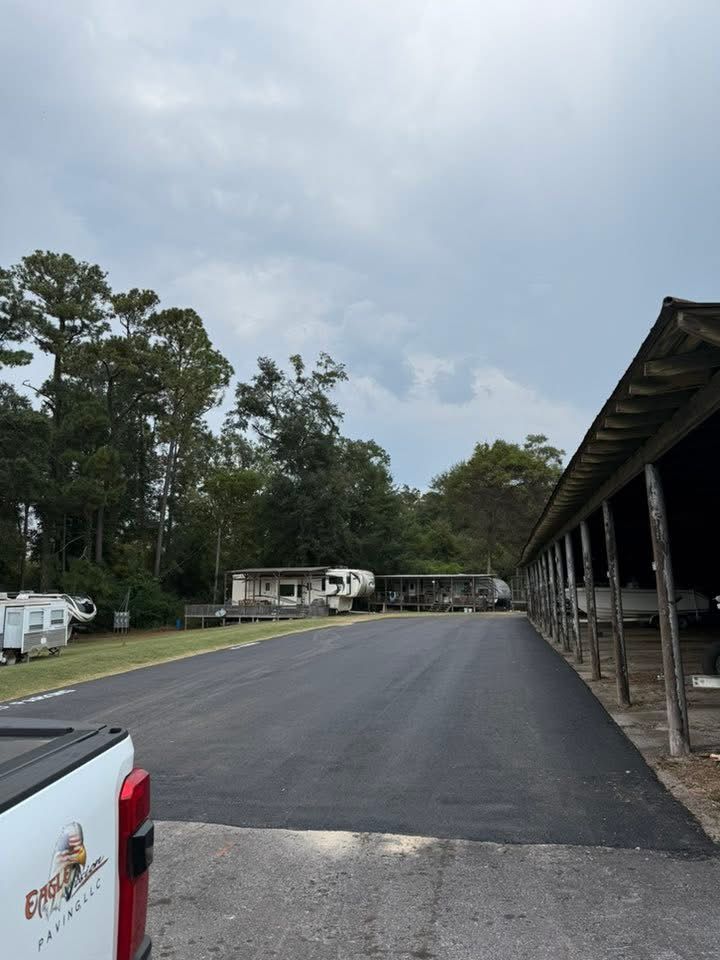 A newly paved asphalt driveway leads toward parked RVs and a wooden shed structure under a cloudy sky.