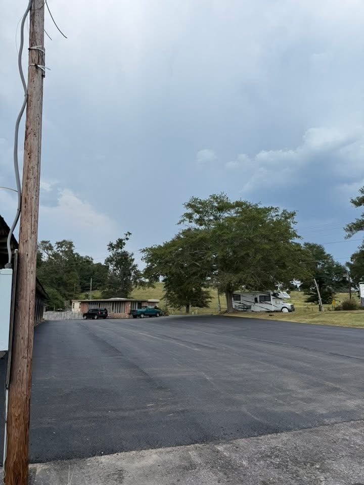 A newly paved black asphalt lot in front of a small house and RV under a cloudy sky.