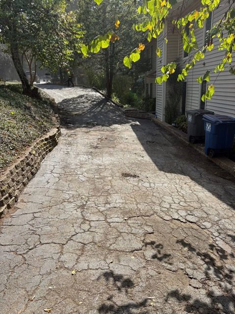 A severely cracked, weathered concrete driveway leading to a house, flanked by a low stone wall and trees.