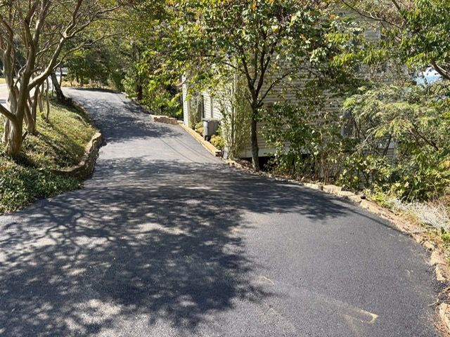 A paved driveway curves uphill toward a partially visible house surrounded by trees on a sunny day.