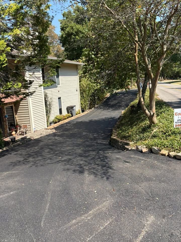 A sloped, paved driveway leads down toward the side of a light-colored multi-story house near trees and a road.