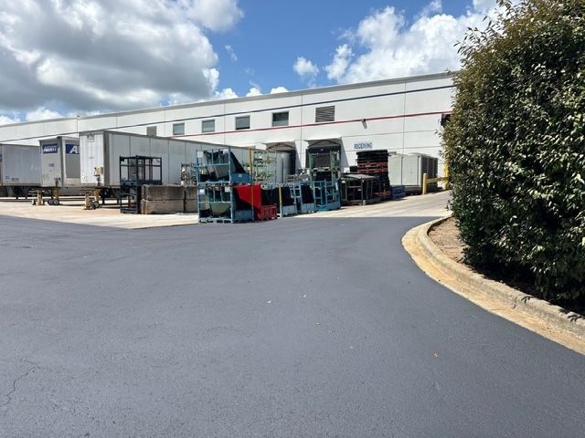 Loading docks at a large white warehouse facility with stacked shipping racks on a paved lot beside a large shrub.