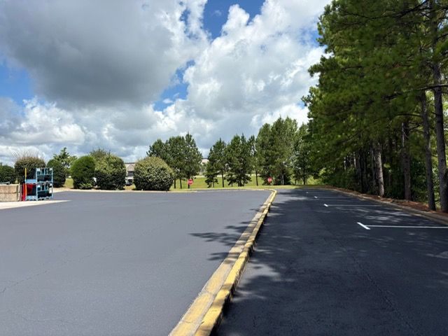 A freshly paved asphalt parking lot on a sunny day with green trees and a blue, cloudy sky in the background.