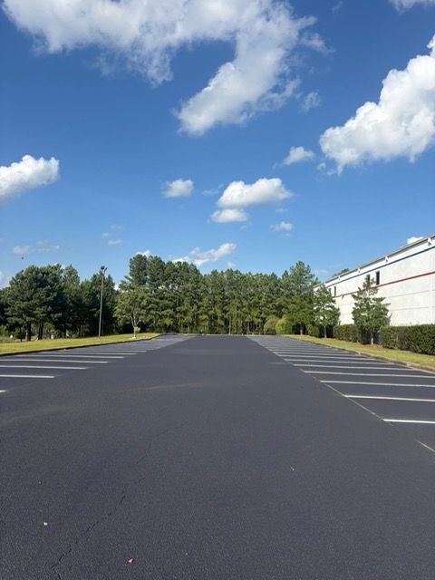 An empty, freshly paved parking lot with painted lines, surrounded by trees under a bright blue sky with white clouds.