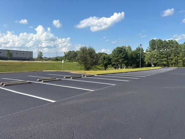 An empty, freshly paved parking lot with painted white spaces and concrete parking stops under a bright, blue sky.