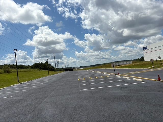 An empty paved parking lot with marked stalls under a bright, partly cloudy sky near a large white building.