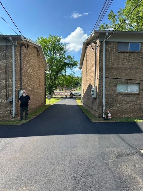 A person stands in a gap between two brick apartment buildings with a freshly paved asphalt driveway extending between them.