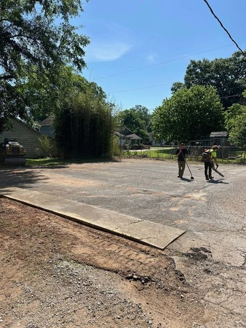 Two people use leaf blowers to clean an outdoor paved lot on a sunny day.