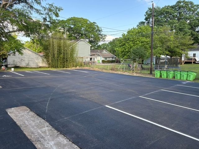 An asphalt parking lot with white painted spaces, a strip of concrete, and a row of green bins near trees and houses.