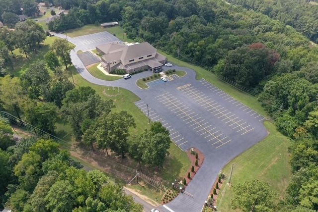 Aerial view of a tan building surrounded by forest, featuring a large parking lot and a circular driveway.