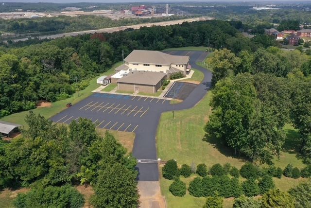Aerial view of a building with a parking lot, surrounded by dense green trees and a paved access road.