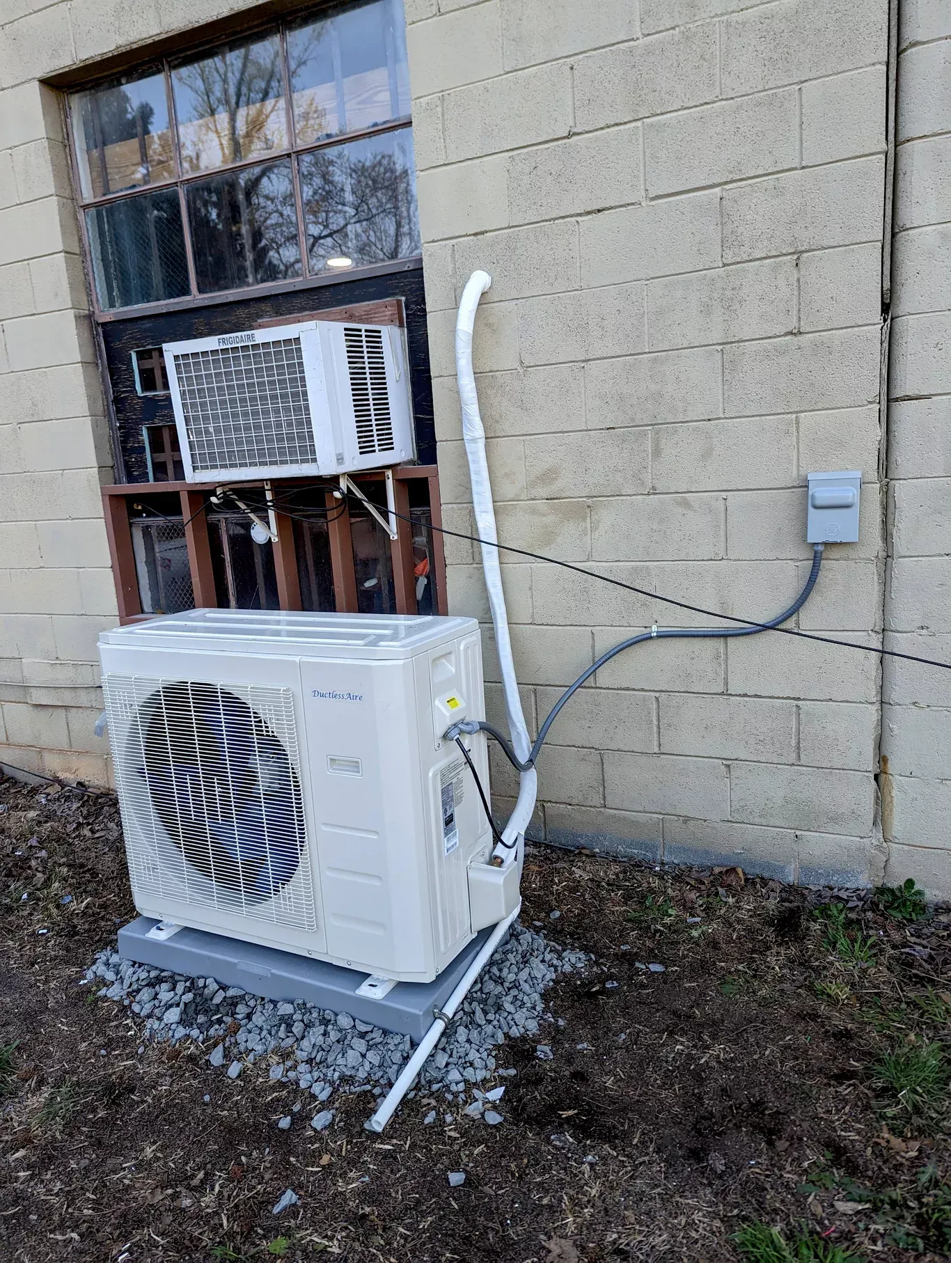 A white air conditioner is sitting outside of a brick building next to a window.