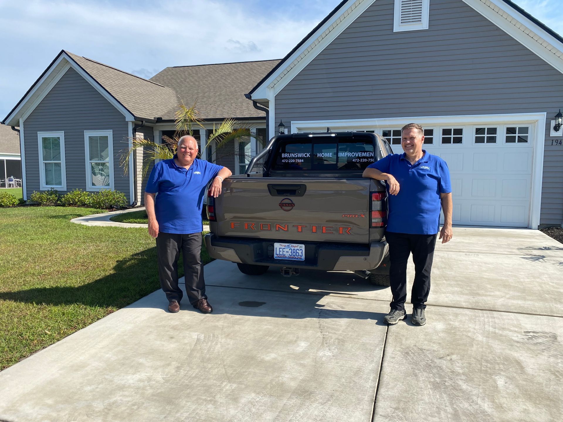 Two people in blue shirts stand beside a brown pickup truck in a driveway in front of a house.