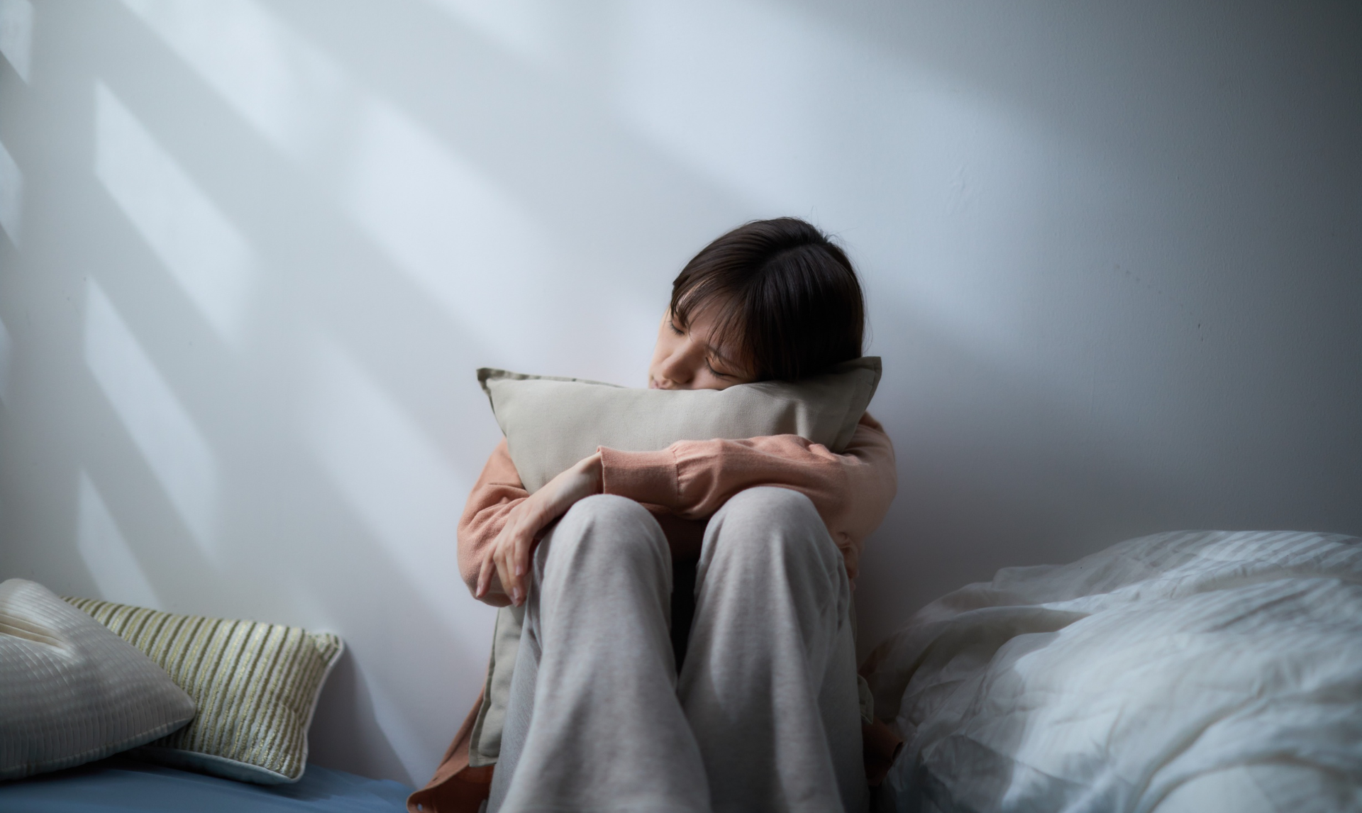 Person seated, hugging pillow, near a wall. Harsh light, shadow stripes. Appears distressed.
