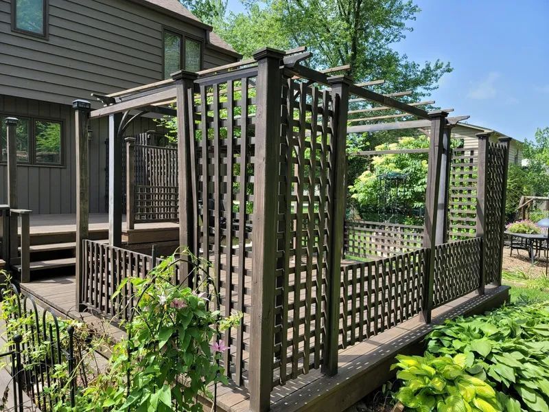 Brown wooden deck with trellis and pergola overlooking a lush green garden.
