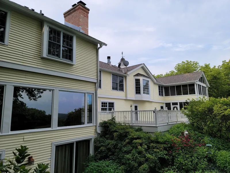 Yellow house with large windows and a deck, surrounded by green foliage under a cloudy sky.
